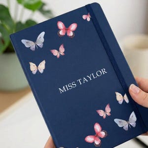 May include: A navy blue hardback notebook featuring a vertical elastic closure band. The cover is decorated with various watercolour-style butterflies in shades of pink, white, and light blue. The text MISS TAYLOR is printed in white capital letters.