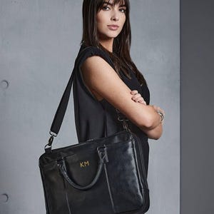 May include: Black leather briefcase with a shoulder strap and top handle. The briefcase features gold-coloured initials "KM" on the front. The bag is carried by a person wearing a black dress.