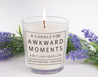 Pode incluir: Uma vela branca acesa num frasco de vidro transparente com um rótulo que diz A CANDLE FOR AWKWARD MOMENTS A BIT LIKE YOUR LIFE With notes of Vanilla & Cookies lizzielane.com/etsy, ladeada por ramos de flores de lavanda roxa.