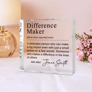 A clear glass block with a definition of a " Difference Maker" written on it, placed on a table with a lamp and a vase of flowers in the background.