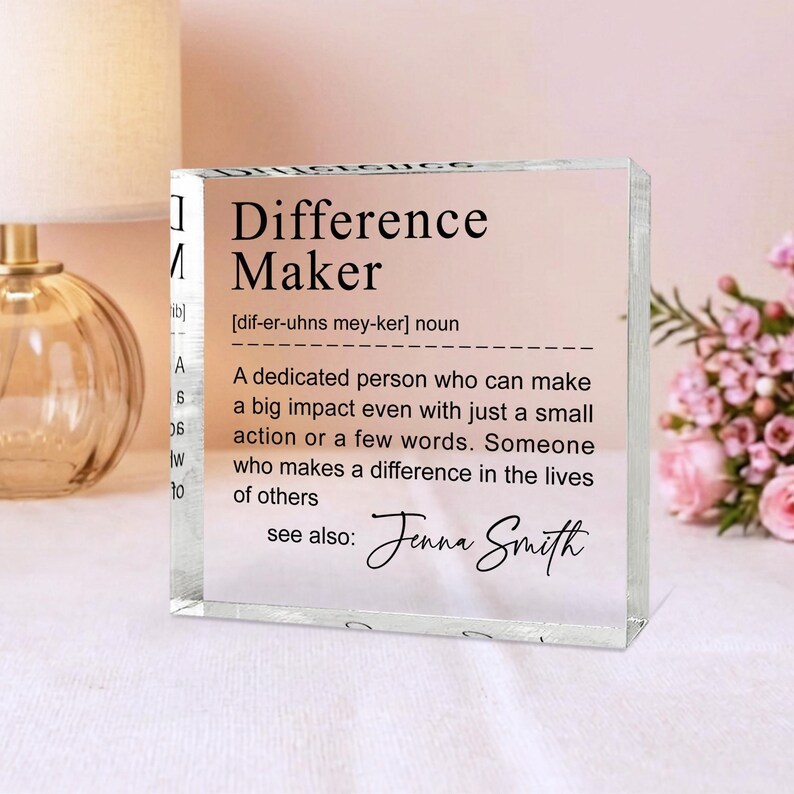 A clear glass block with a definition of a " Difference Maker" written on it, placed on a table with a lamp and a vase of flowers in the background.