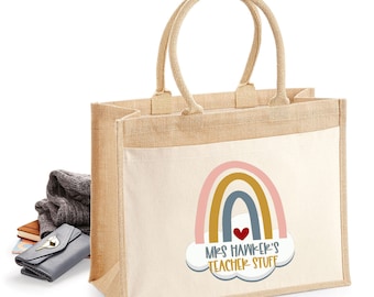 May include: A rectangular jute tote bag with two top handles and a white front pocket. The pocket features a printed graphic of a rainbow in muted tones, a small red heart, and the text "MRS HAWKER'S TEACHER STUFF" inside a white cloud shape.
