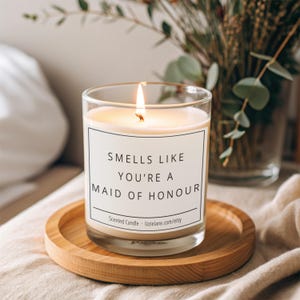 Könnte beinhalten: Eine weiße Kerze mit einem schwarzen Etikett, auf dem "Smells like you're a Maid of Honour" steht. Die Kerze ist angezündet und steht auf einem Holzuntersetzer.