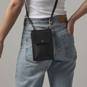 A person wearing a white shirt and blue jeans, with a black crossbody bag featuring a gold chain strap and a personalized logo.