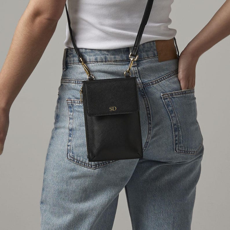 A person wearing a white shirt and blue jeans, with a black crossbody bag featuring a gold chain strap and a personalized logo.