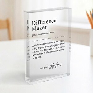 A clear glass block with a definition of " Difference Maker" written on it, placed on a table with a white surface and a white mug in the background.