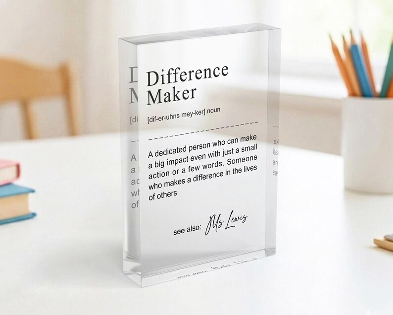 A clear glass block with a definition of " Difference Maker" written on it, placed on a table with a white surface and a white mug in the background.