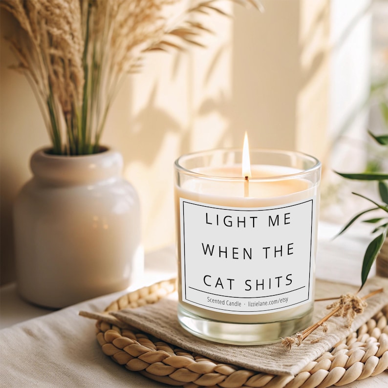 May include: A white candle with a black label that reads "Light me when the cat shits". The candle is lit and sitting on a woven placemat.