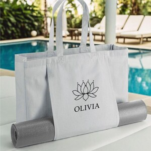 A white tote bag with a black logo and text, placed on a gray mat, with a pool and palm trees in the background.