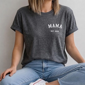 Peut inclure: Un t-shirt gris chiné foncé à manches courtes avec des revers. L'avant présente un texte blanc sur la poitrine gauche indiquant MAMA dans une police serif grasse, avec le texte EST 2026 imprimé en plus petit juste en dessous.