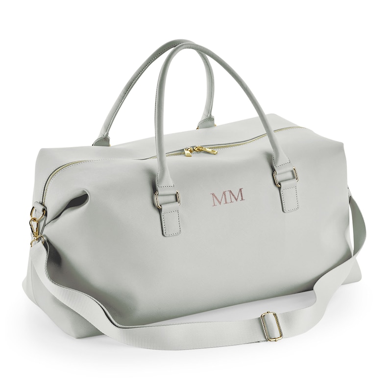 May include: A light gray leather duffel bag with a gold zipper and gold hardware. The bag has a long shoulder strap and two top handles. The initials "MM" are embroidered on the front of the bag.