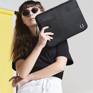 Könnte beinhalten: Eine schwarze, rechteckige Laptoptasche mit Reißverschluss und den weiß gestickten Initialen LJ in der unteren rechten Ecke. Die Tasche hat eine glatte, stoffartige Textur und ein schmales Profil für den Transport eines Laptops.
