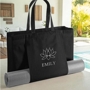 A black tote bag with a white lotus flower design and the name "EMILY" printed on it, placed on top of a gray yoga mat.