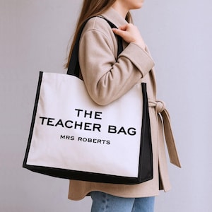 A woman wearing a beige coat is carrying a large white bag with black text that says "THE TEACHER BAG MRS ROBERTS".