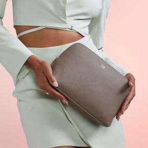 May include: A taupe-colored rectangular zippered pouch with a textured finish. The front of the pouch features the letters TB embossed in a small, centered font. The pouch is held in place, showcasing its clean lines and minimalist design.