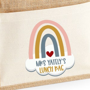 May include: A beige canvas lunch bag with a rainbow design. The rainbow is composed of pink, gold, and blue arches, with a red heart and the text "MRS YATELY'S LUNCH BAG" in a playful font. The bag has a burlap trim.