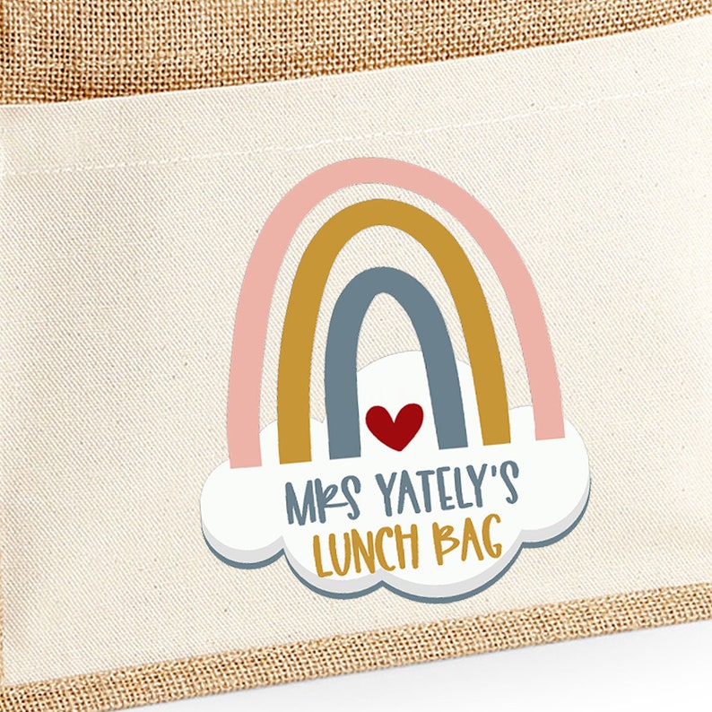 May include: A beige canvas lunch bag with a rainbow design. The rainbow is composed of pink, gold, and blue arches, with a red heart and the text "MRS YATELY'S LUNCH BAG" in a playful font. The bag has a burlap trim.
