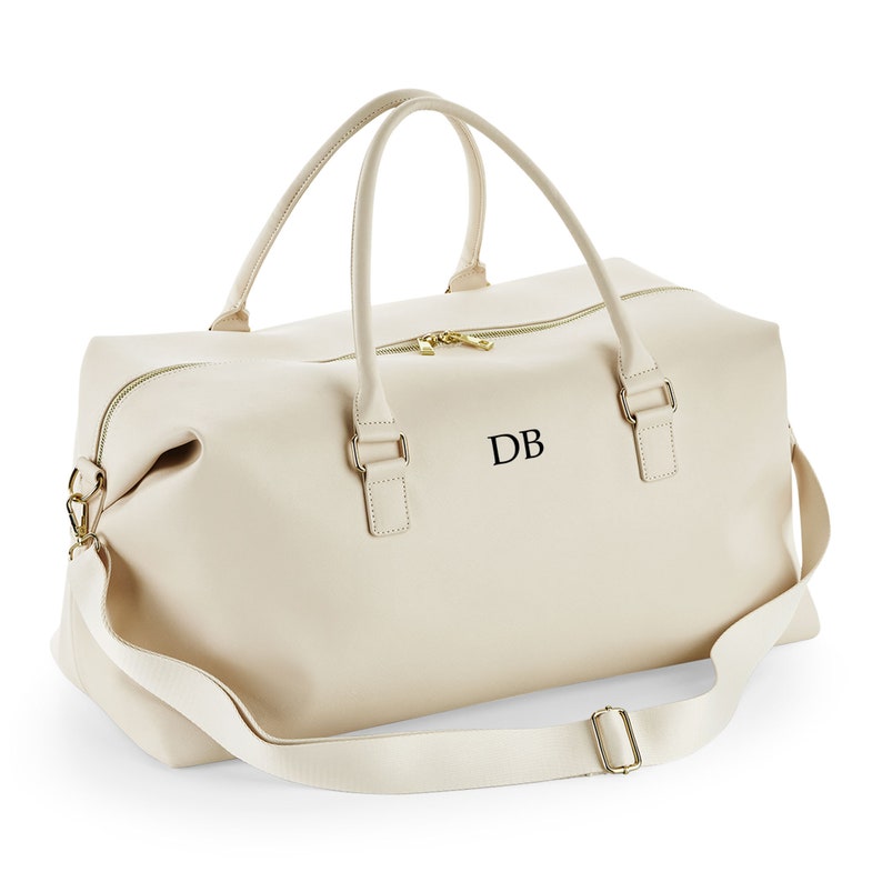 May include: A cream-colored leather duffel bag with a gold zipper and a black leather strap. The bag has a gold buckle and the initials "DB" are embroidered on the front.