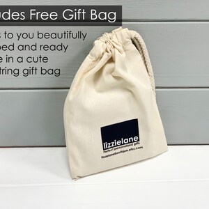 A beige drawstring gift bag with a black logo and text, placed on a white surface against a gray background.