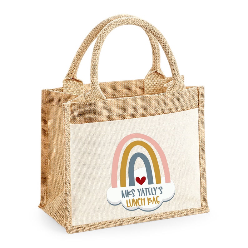 May include: A beige burlap lunch bag with a white pocket featuring a rainbow design and the text "Mrs. Yately's Lunch Bag". The bag has two handles and is designed for carrying lunches.