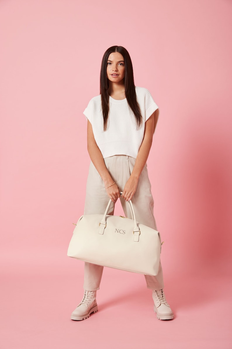 May include: A white leather travel bag with gold hardware and the initials "NCS" embossed on the front. The bag is being held by a person wearing a white sweater and beige pants.