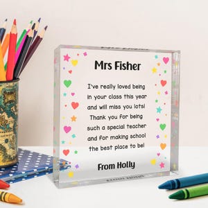 Peut inclure: Un bloc acrylique transparent avec le texte "Mrs Fisher" et un message sincère, décoré de cœurs et d'étoiles colorés. Le message est un remerciement de "Holly". Un pot à crayons et des crayons sont en arrière-plan.