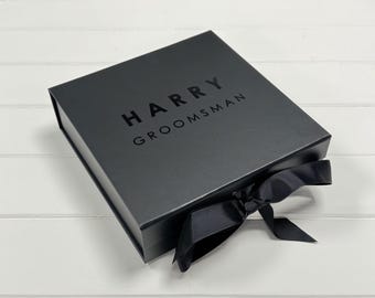 May include: A square black gift box with a hinged lid, featuring the text HARRY GROOMSMAN printed in black on the top surface. The box is secured with a black satin ribbon tied into a bow at the front edge, creating a sleek and uniform appearance.
