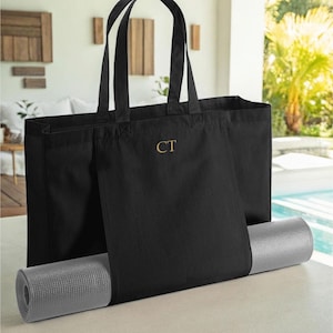 May include: Black canvas tote bag with a pocket for a yoga mat. The bag has a gold monogram "CT" on the front.