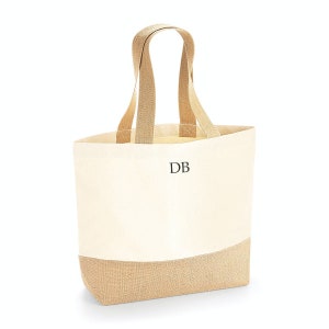 Pu&ograve; includere: Una borsa tote in tela naturale con dettagli in juta e manici in tessuto marrone. La borsa ha una sezione superiore color crema e una sezione inferiore in juta marrone. La borsa ha le iniziali "DB" ricamate sul davanti.