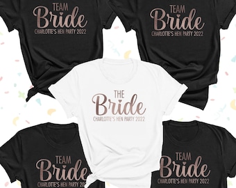 May include: Four black t-shirts and one white t-shirt with rose gold lettering. The black shirts feature the text TEAM Bride CHARLOTTE'S HEN PARTY 2022. The white shirt features the text THE Bride CHARLOTTE'S HEN PARTY 2022. All shirts have short sleeves.