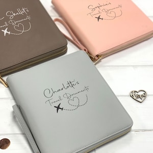 May include: Three rectangular zippered travel document organizers in brown, light pink, and light gray. Each features a wrist strap, gold-tone zipper, and custom text: The Skellet's Travel Documents, Sophie's Travel Documents, and Charlotte's Travel Documents.