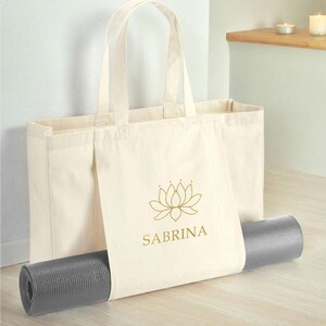 A beige tote bag with the word "SABRINA" printed on it, along with a gray yoga mat, is placed on a wooden floor.