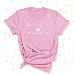 A pink t-shirt with the text "Bridal Squad Charlotte" printed on it, along with a heart symbol.