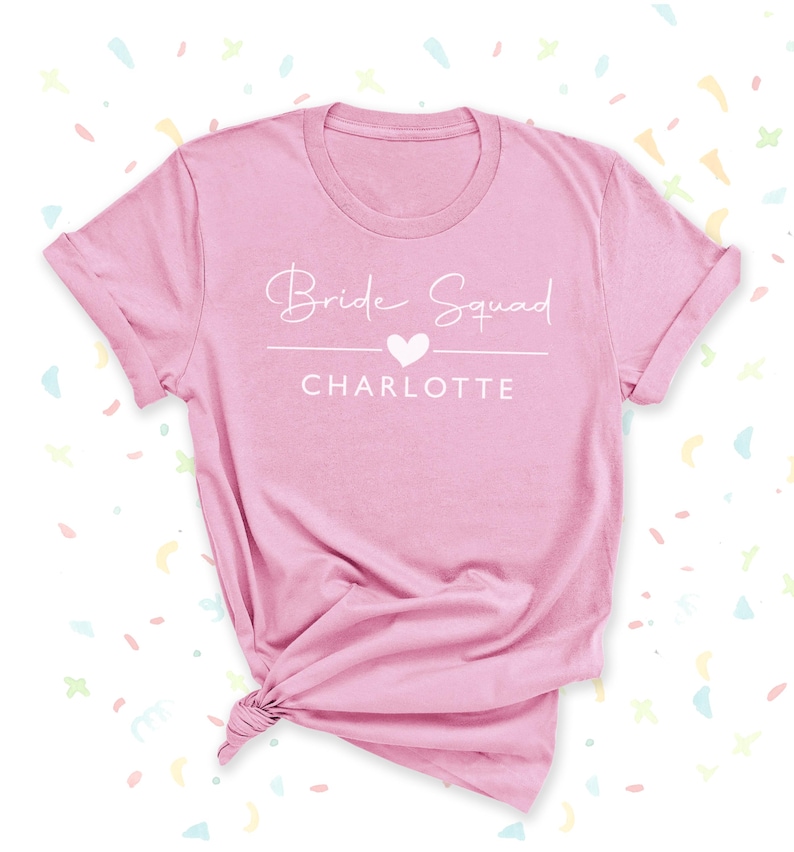 A pink t-shirt with the text "Bridal Squad Charlotte" printed on it, along with a heart symbol.