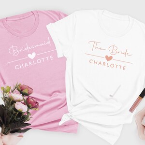 two t-shirts, one pink and one white, with the text "Bridesmaid" and "The Bride" printed on them. There is also a bouquet of flowers and a small bowl of white powder or sugar cubes on the side.