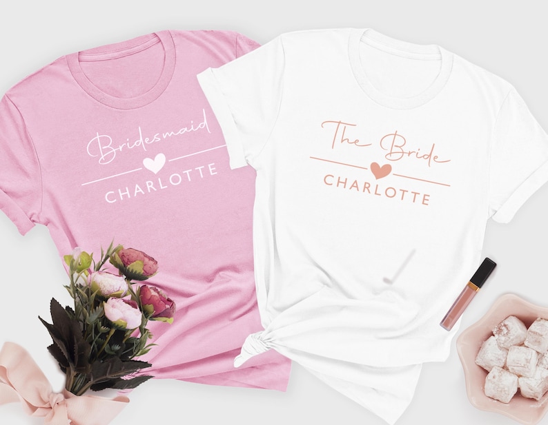 two t-shirts, one pink and one white, with the text "Bridesmaid" and "The Bride" printed on them. There is also a bouquet of flowers and a small bowl of white powder or sugar cubes on the side.