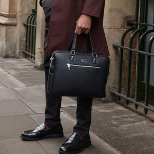May include: A black leather briefcase with silver zippers and handles, held by a person. The briefcase has the initials "MG" on the front. The person is wearing black pants and shiny black loafers.