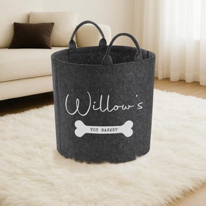 A gray felt toy basket with a white bone-shaped handle, labeled "Willow's Toy Basket" in white text, sits on a white shag rug in a living room.