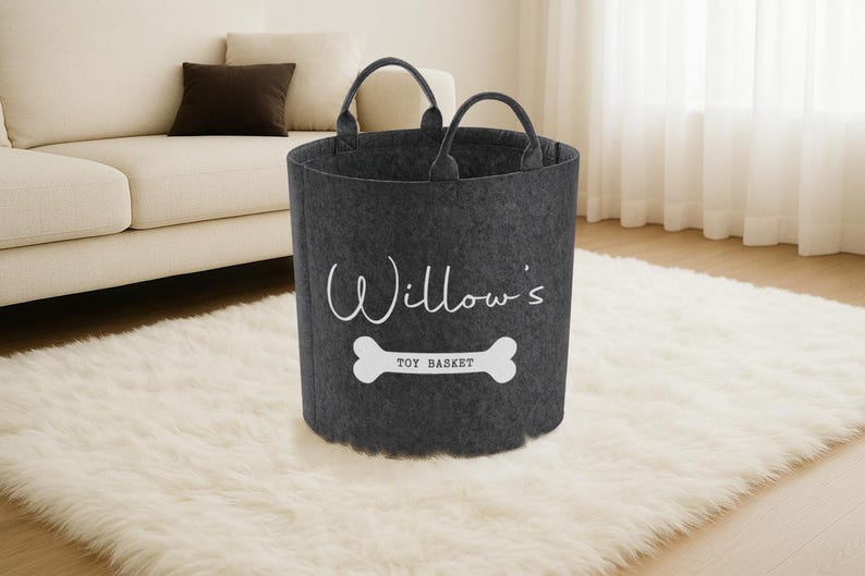 A gray felt toy basket with a white bone-shaped handle, labeled "Willow's Toy Basket" in white text, sits on a white shag rug in a living room.