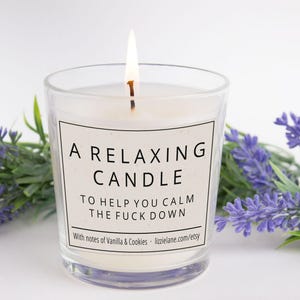 May include: A lit white candle in a clear glass jar with a label that reads A RELAXING CANDLE TO HELP YOU CALM THE FUCK DOWN with notes of Vanilla & Cookies lizzielane.com/etsy, surrounded by sprigs of purple lavender flowers.