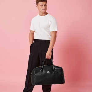 May include: A black leather travel bag with gold hardware and the initials "LR" embroidered on the front. The bag is being held by a person wearing a white t-shirt and black pants.
