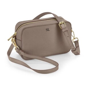 May include: A taupe leather crossbody bag with a gold zipper and buckle. The bag has a short strap and a long adjustable strap. The initials "SL" are embossed on the front of the bag.
