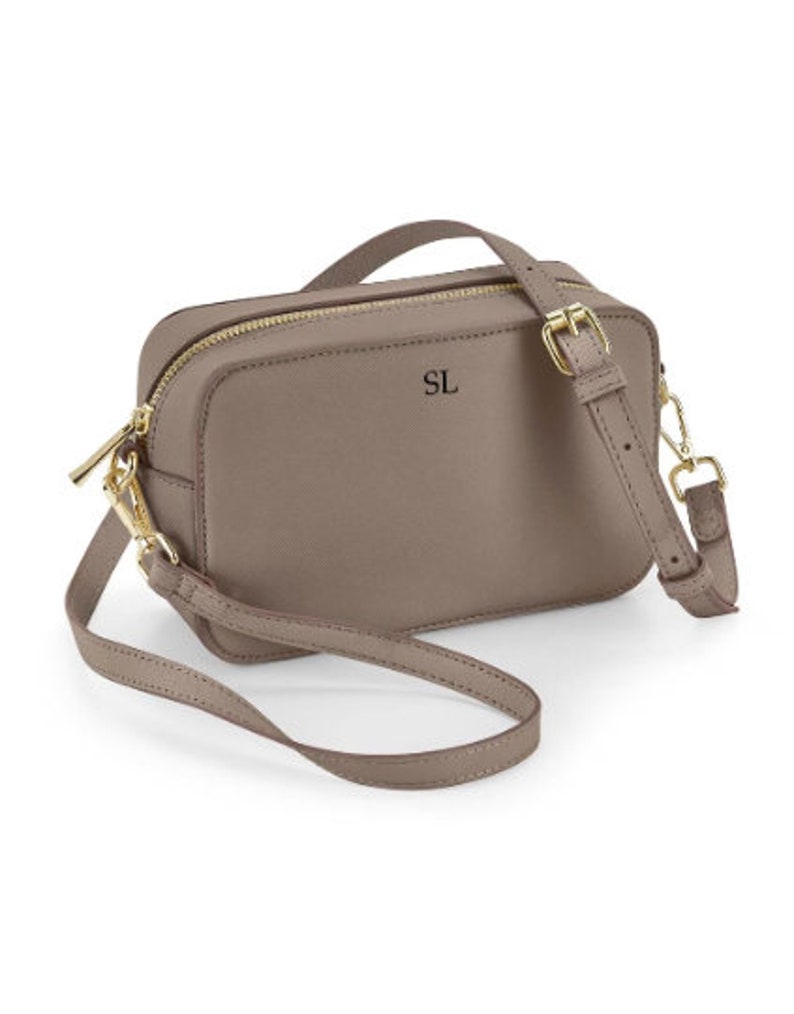 May include: A taupe leather crossbody bag with a gold zipper and buckle. The bag has a short strap and a long adjustable strap. The initials "SL" are embossed on the front of the bag.