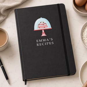 May include: A black hardcover recipe journal with an elastic closure band. The front cover features a graphic of a pink cake on a stand under a glass dome, with the white text EMMA'S RECIPES printed directly below the illustration.