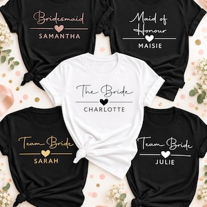 Könnte beinhalten: Fünf kurzärmelige T-Shirts mit Rundhalsausschnitt. Vier sind schwarz mit weißem, roségoldenem oder goldenem Text und Herzsymbolen mit der Aufschrift Bridesmaid Samantha, Maid of Honour Maisie, Team Bride Sarah und Team Bride Julie. Ein weißes Shirt zeigt The Bride Charlotte.