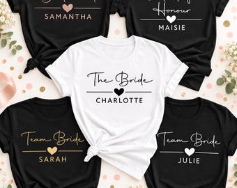 May include: Five short-sleeved crew neck t-shirts. Four are black with white, rose gold, or gold text and heart icons reading Bridesmaid Samantha, Maid of Honour Maisie, Team Bride Sarah, and Team Bride Julie. One white shirt reads The Bride Charlotte.