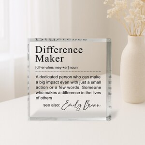 A clear glass block with a definition of a " Difference Maker" written on it, along with a quote from Emily Brown. The block is placed on a table, and there is a vase of flowers in the background.