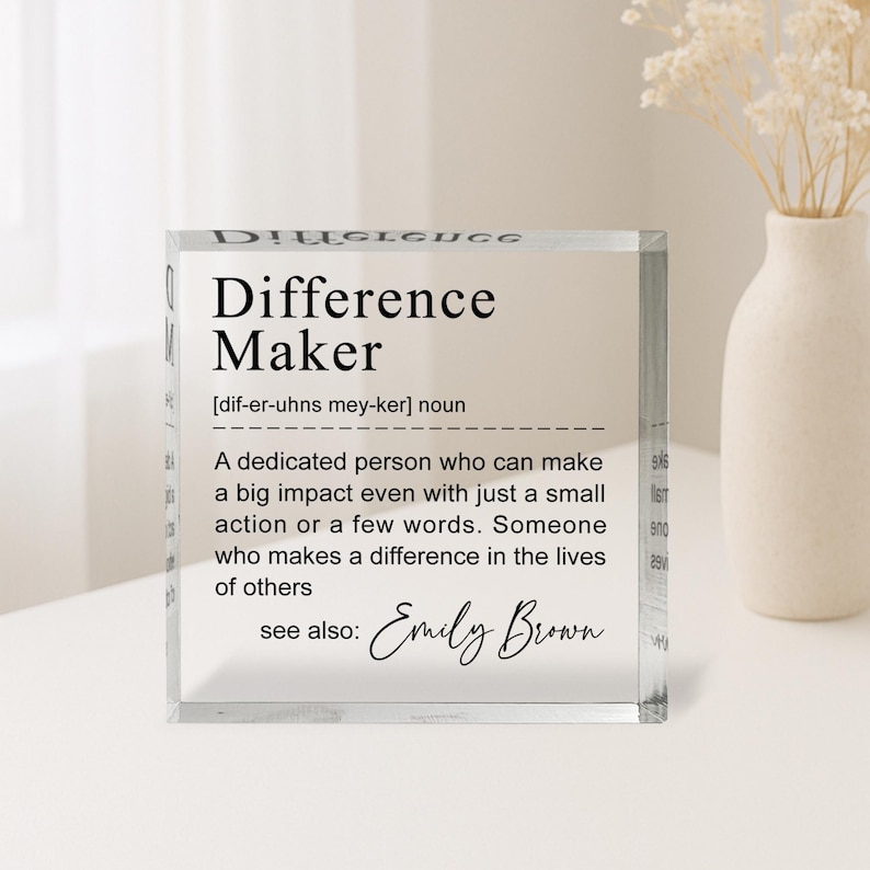 A clear glass block with a definition of a " Difference Maker" written on it, along with a quote from Emily Brown. The block is placed on a table, and there is a vase of flowers in the background.
