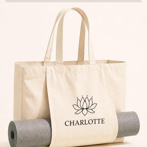 A beige canvas tote bag with a printed logo and the name "CHARLOTTE" printed on it, along with a gray yoga mat rolled up inside the bag.