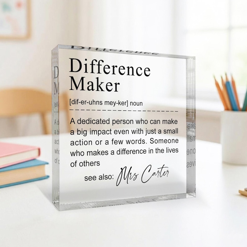A clear glass block with a definition of a " Difference Maker" written on it, placed on a desk with books and a white mug in the background.